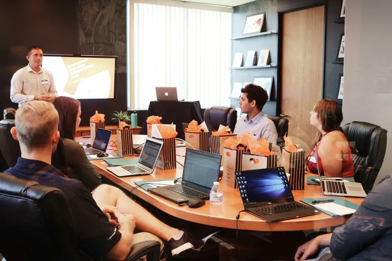 A presenter in front of a team seated around a table with laptops